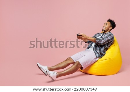 Full body young fun man of African American ethnicity wear blue shirt sit in bag chair hold play pc game with joystick console isolated on plain pastel light pink background. People lifestyle concept