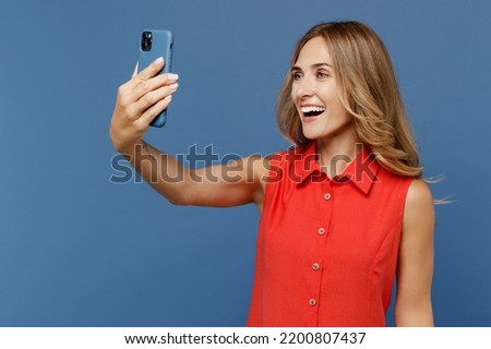 Young smiling woman she wear red dress doing selfie shot on mobile cell phone post photo on social network isolated on plain dark royal navy blue background studio portrait. People lifestyle concept
