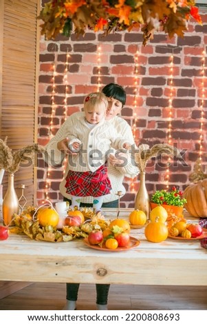 A young mother and little daughter in white warm knitted sweaters in an interior decorated with blankets and pillows
 as well as pumpkins, autumn leaves and apples. Autumn mood. halloween