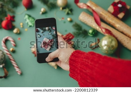 Hand taking photo of stylish wrapped christmas gift, golden paper, red ribbon and festive decorations on green background. Social media. Merry Christmas and Happy Holidays!