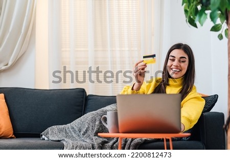 Delighted young brunette woman, holding up her credit card, checking her bank account from bed on laptop.