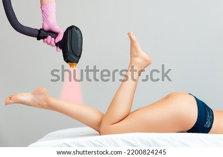 Cosmetologists hand in medical glove holds a laser hair removal device. The laser beam is aimed at woman's slender legs. Concept of hair removal and apparatus cosmetology. Royalty-Free Stock Photo #2200824245