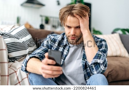 Frustrated unhappy caucasian young stylish man, sitting on a floor near a sofa in living room, sadly looking at smartphone screen, feel loneliness, got bad news or message, despair emotions Royalty-Free Stock Photo #2200824461