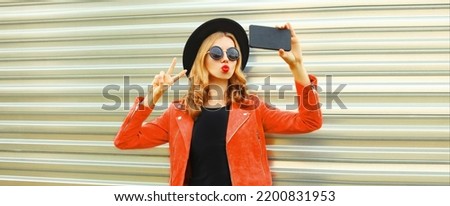 Portrait of beautiful happy woman taking selfie with smartphone and blowing her lips sends air kiss wearing black round hat, red leather jacket on gray background
