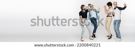 young woman showing victory sign and dancing near friends taking photo on smartphones on grey background, banner