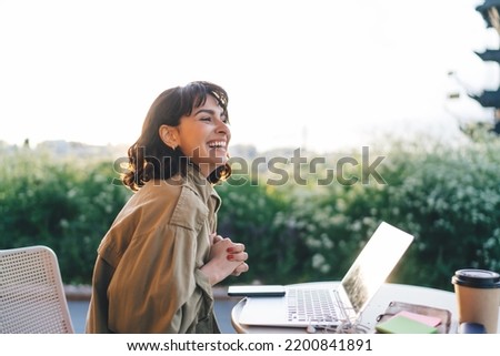 Side view of young brunette smiling happily while sitting at table with laptop working remotely on new web design project in sunny garden Royalty-Free Stock Photo #2200841891