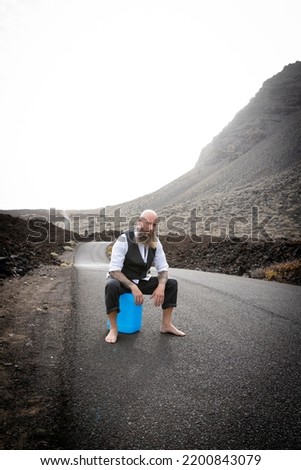 Man with suit is sitting completely desperate and knocked out on a blue gasoline canister somewhere in the nowhere in the volcanic desert at the end of the world and is not happy