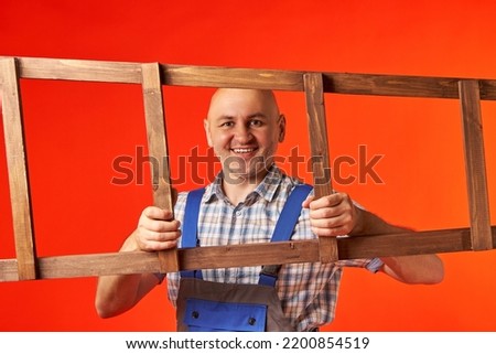 Bald man in a work suit smiles between the windows of a wooden ladder, which he holds horizontally, on a red background