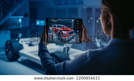 Automotive Engineer Working on Electric Car Chassis Platform, Using Tablet Computer with Augmented Reality 3D Software. Innovative Facility: Vehicle Frame with Wheels Becomes a VFX Virtual Model. Royalty-Free Stock Photo #2200856615