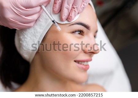 Close up of hands of young cosmetologist injecting botox in female face. She is standing and smiling. The woman is closed her eyes with relaxation Royalty-Free Stock Photo #2200857215