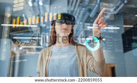 Young Adult Female Using Futuristic Augmented Reality Application Interface for Managing Business and Marketing Projects. Professional in Office Wearing Headset to Look at VFX Edit with Financial Data Royalty-Free Stock Photo #2200859055