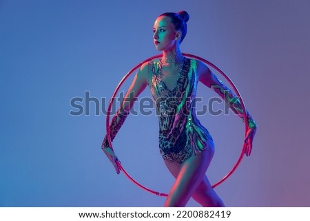 Portrait of young, muscular girl, female rhytmic gymnast training with hoop isolated over blue studio background in neon light. Concept of action, motion, sport life, motivation, competition.