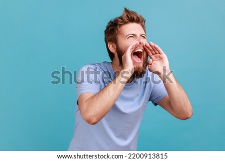 Portrait of angry handsome bearded man loudly screaming widely opening mouth holding hands near mouth, yelling announcing his opinion. Indoor studio shot isolated on blue background. Royalty-Free Stock Photo #2200913815