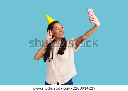 Happy woman in cone broadcasting livestream, waving hand to followers, recording video for blog during celebration her birthday, wearing white shirt. Indoor studio shot isolated on blue background.