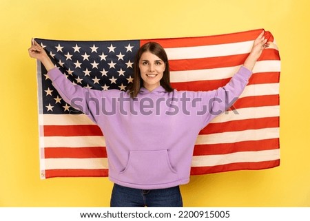Portrait of attractive happy positive woman standing with raised arms, holding USA flag, celebrating national holiday, wearing purple hoodie. Indoor studio shot isolated on yellow background.