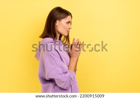 Side view of devious cunning young woman clasping hands and smirking mysteriously, scheming cheats, evil prank, wearing purple hoodie. Indoor studio shot isolated on yellow background. Royalty-Free Stock Photo #2200915009