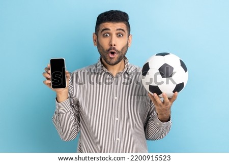 Surprised bearded man holding soccer ball and smartphone empty black display, shocked with easy championship ticket booking, wearing striped shirt. Indoor studio shot isolated on blue background.