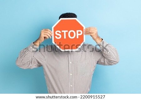 Portrait of man covering face with Stop symbol, anonymous person holding red traffic sign, warning to go, prohibition concept, wearing striped shirt. Indoor studio shot isolated on blue background.