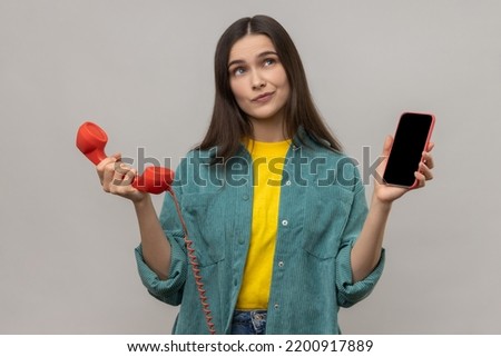 Portrait of confused puzzled woman standing retro telephone and mobile phone, thinking what to use better, wearing casual style jacket. Indoor studio shot isolated on gray background. Royalty-Free Stock Photo #2200917889