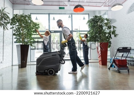 side view of man in overalls with electrical floor scrubber machine near interracial colleagues Royalty-Free Stock Photo #2200921627