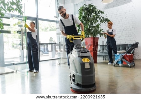 man in workwear with floor scrubber machine near interracial women cleaning office lobby Royalty-Free Stock Photo #2200921635