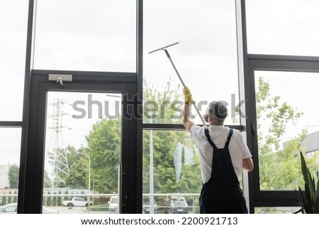 back view of man in overalls washing large office windows with window squeegee Royalty-Free Stock Photo #2200921713