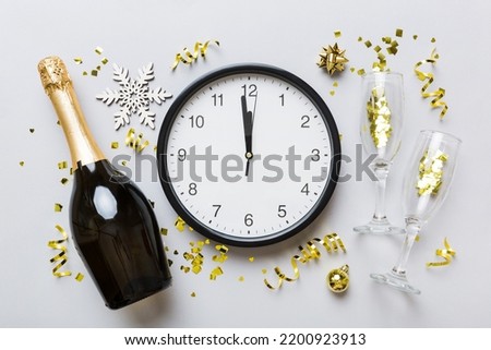 New year celebration concept with a bottle of champagne and two glasses toasting. Christmas gift box, alarm clock and fir tree branch on colored table. Top view Copy space.