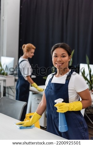 happy bi-racial woman in overalls and rubber gloves smiling at camera near blurred colleague