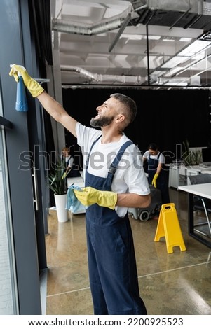 bearded man in workwear cleaning window with detergent near multiethnic women working in office
