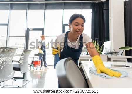 smiling bi-racial woman cleaning office desk with rag near colleague on blurred background Royalty-Free Stock Photo #2200925613