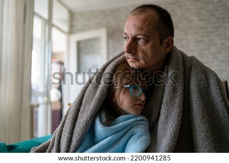 Dissatisfied father and daughter having problem with central heating, sitting on sofa at home, freezing. Freezing family warm blankets looking at camera while sitting on sofa in cold kitchen Royalty-Free Stock Photo #2200941285