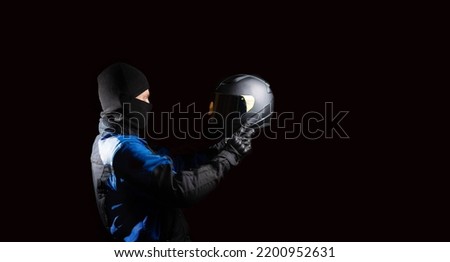 male motorcyclist in a motorcycle outfit and a balaclava puts on a helmet on a dark background. The concept of protective equipment for a biker. Royalty-Free Stock Photo #2200952631