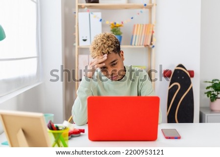 Stressed teenager boy sitting at desk studying on laptop in room. Hispanic latin young student man worried about school exams.