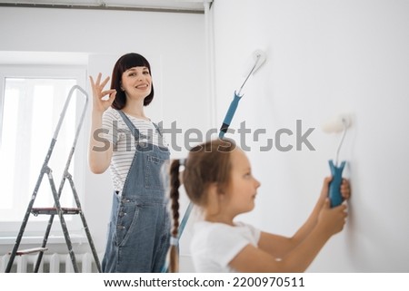 Young family of beautiful mother and cute little daughter having fun together while repairing room. Little girl with paint roller coloring with mom the wall. Focus on mum showing sign ok.