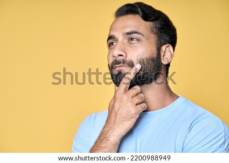 Thoughtful bearded indian man holding hand on chin looking interested aside at copy space isolated on yellow background thinking of new job opportunities, having doubt question or deciding concept. Royalty-Free Stock Photo #2200988949