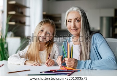 Cheerful caucasian little girl with aged lady with colorful pencils draw a picture on table, enjoy study and free time in living room interior. Art lesson together, education and entertainment at home