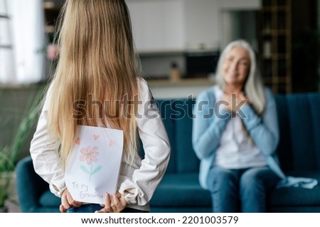 European small granddaughter holds behind back postcard with picture of happy senior female in living room interior. Surprise birthday greeting and holiday celebration at home, love and relationship