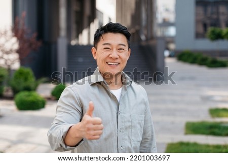 Portrait of positive asian man standing outdoors and gesturing thumb up, walking in urban city area, looking and smiling at camera, free copy space Royalty-Free Stock Photo #2201003697