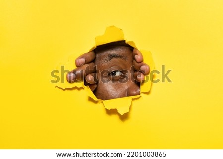 Young african american man peeking out of a hole in yellow paper background, look through torn hole with one eye, free copy space Royalty-Free Stock Photo #2201003865
