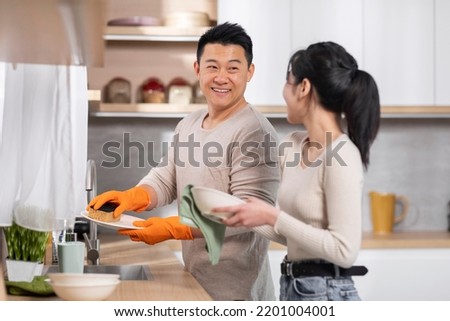 Cheerful asian spouses doing chores together, husband washing dishes and wife drying up plates, happy couple having conversation and smiling, side view, copy space