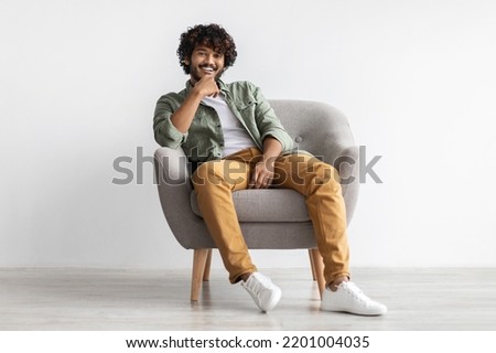 Relaxed handsome young indian guy in stylish casual outwear chilling in arm chair over white background, holding hand on his chin and laughing at camera, full length, copy space