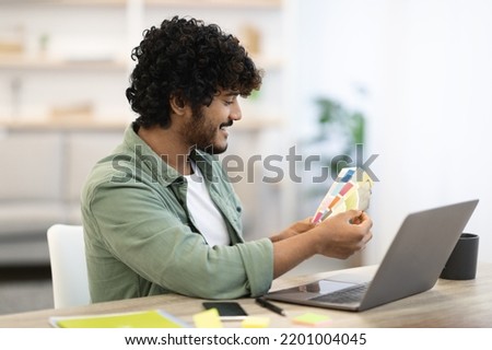 Cheerful dark-skinned young guy with curly hair sitting at workdesk in front of laptop at home office, checking color palettes, indian man web-designer making website, copy space