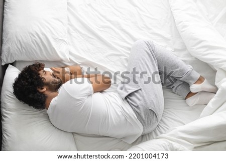 Upset millennial hindu guy in white pajamas lying on bed at home, feeling down, experiencing troubles, top view, looking at empty bed space by him. Depression, loneliness concept Royalty-Free Stock Photo #2201004173