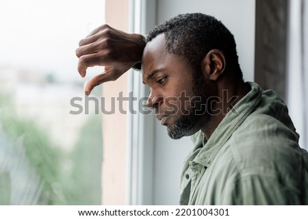 Unhappy frustrated adult african american guy in casual suffering from depression and bad news near window in home interior. Health problems, stress from self-isolation during covid-19 quarantine Royalty-Free Stock Photo #2201004301