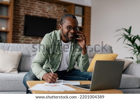 Smiling busy mature african american guy manager in glasses and casual, have call by phone, works on laptop in living room interior. Business remotely, freelance, new normal with device and meeting Royalty-Free Stock Photo #2201004389