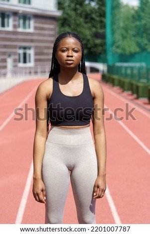 African American sportswoman prepares for running on jogging track at sunlight. Black woman wearing black top and grey leggings stands in powerful posture showing sporty body against blurry racetrack