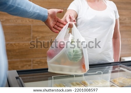 Female saleswoman hands plastic bag with fresh vegetables from hand to hand. Man is shopping in grocery store. Close up of modern trendy biodegradable package used in store. Royalty-Free Stock Photo #2201008597