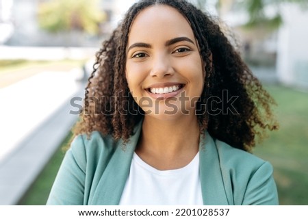 Close-up photo of beautiful positive confident successful hispanic or brazilian young woman with curly hair, business lady, in stylish elegant clothes, standing outdoors, looking at the camera, smiles