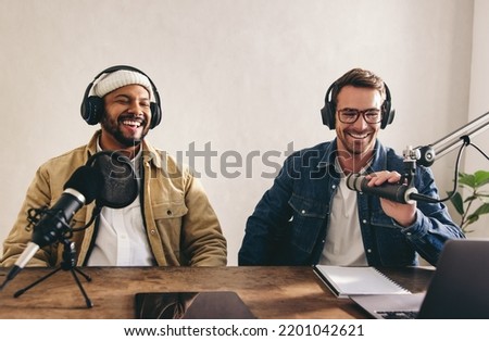 Male radio presenters having a great time on a live show. Two men smiling happily while recording an audio broadcast in a studio. College content creators co-hosting an internet podcast.