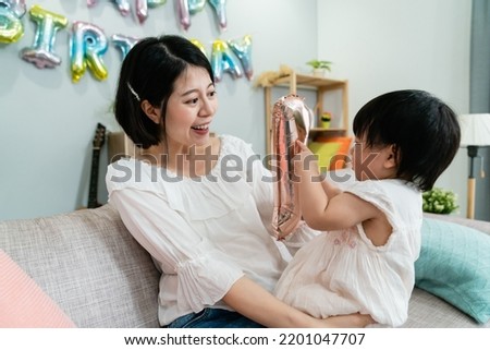 pretty asian mother holding and smiling at her cute baby girl who’s having fun with a number standee on the couch at an indoor birthday party